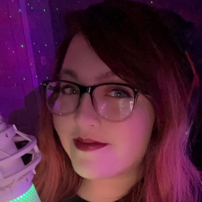 A photograph of Grace. A person with long red hair, light skin tone, glasses with black rims and a smile on their face. The photo is taken in a purple/pink lit room.